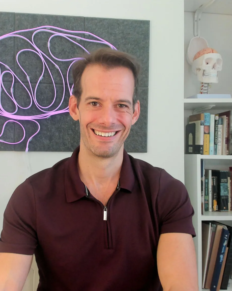 Michael Corayer smiling in front of a neon brain sign and his psychology bookshelf